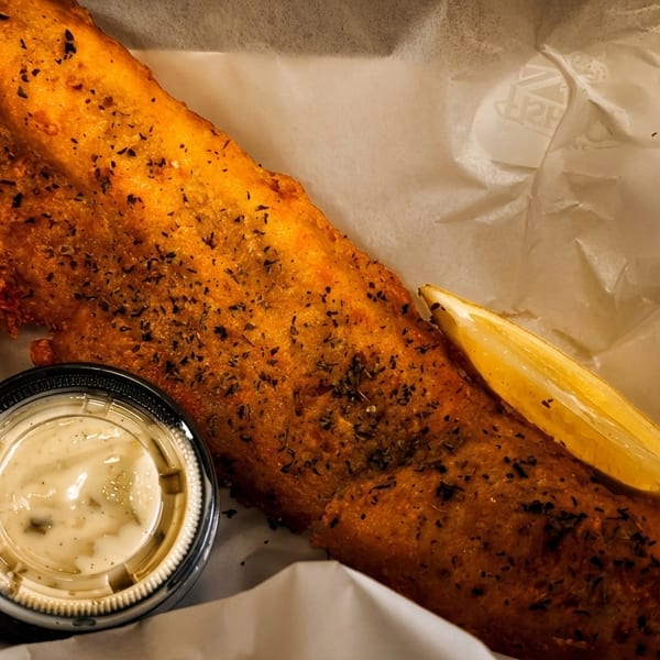Golden-brown fried fish with herbs, a lemon wedge, and tartar sauce in a paper-lined basket.
