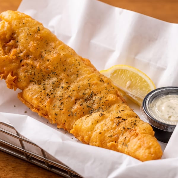 Golden-brown fried fish with herbs, a lemon wedge, and tartar sauce in a metal basket.